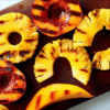 Close-up of colorful leftover grilled fruit, including pineapple, peaches, and mango, arranged on a dark wooden cutting board. The grill marks are visible and appetizing.